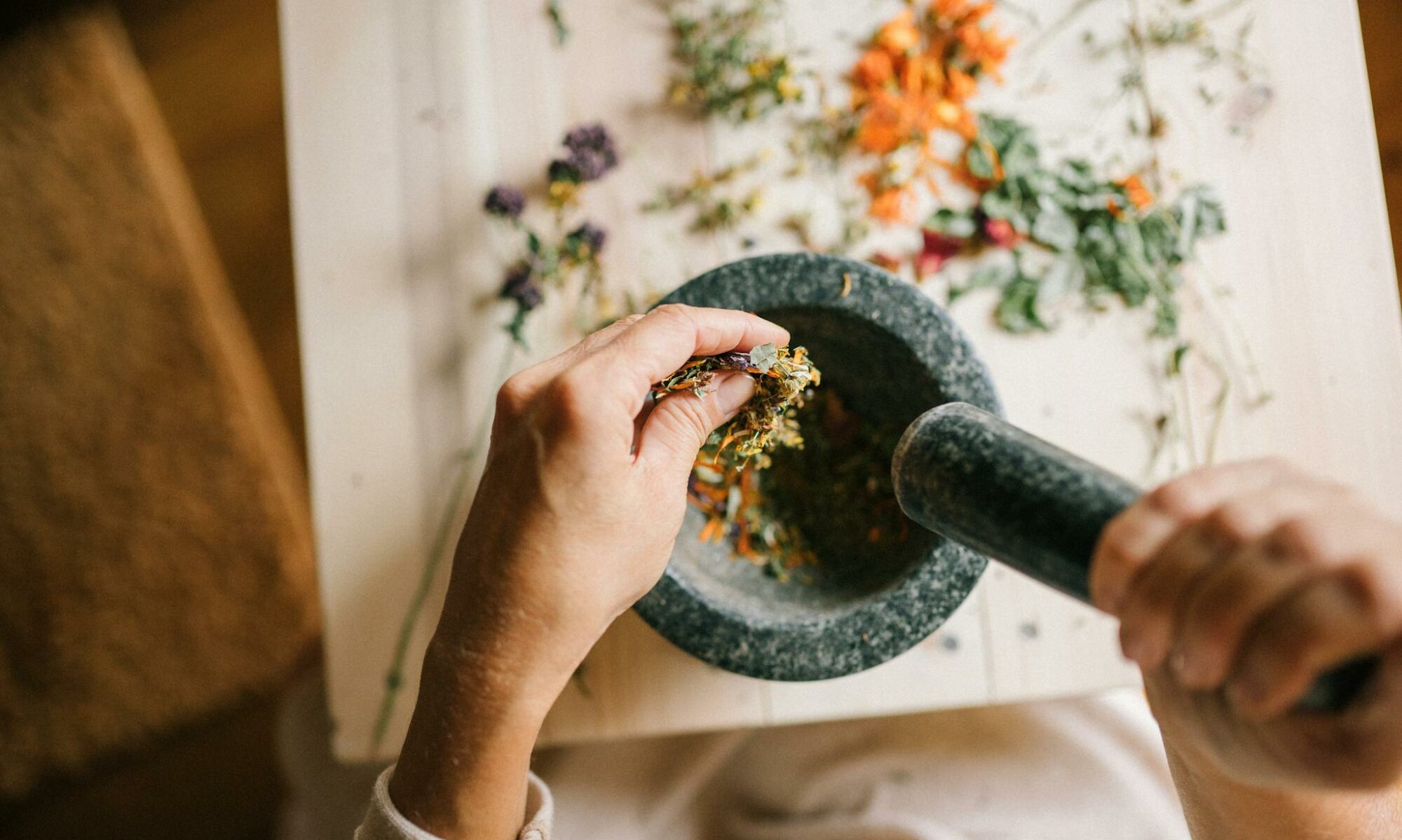 Top view of hands grinding dried herbs using a mortar and pestle on a wooden table.