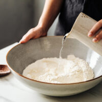 Midsection of woman pouring water on rye and wheat flour in bowl. Female is standing in kitchen. She is preparing sourdough bread.